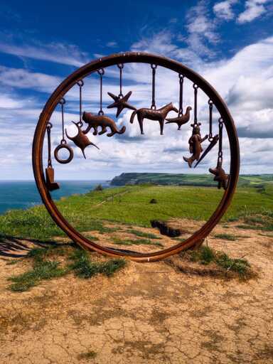 A large circular steel sculpture stands on the clifftop beside the Cleveland Way, framing a sweeping view of green coastal fields, distant sea cliffs and the North Sea beyond. Known locally as the “Charm Bracelet”, the artwork consists of a rust-coloured metal ring from which a collection of hanging shapes is suspended, including a cat, a horse, a bird, starfish-like forms, geometric symbols and a small human figure. The sculpture dominates the foreground, creating a natural frame for the landscape behind, while dramatic white clouds drift across a deep blue sky, enhancing the artwork’s mystical and whimsical character.